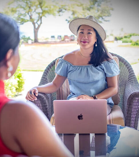 Kris Chau, Small business consultant sits in a wicker chair across from a client, who is only slightly visible with her back and red shirt. Kris is wearing a straw fedora, pearl dangling earrings, and a off the shoulder chambray top and white jeans. In fro