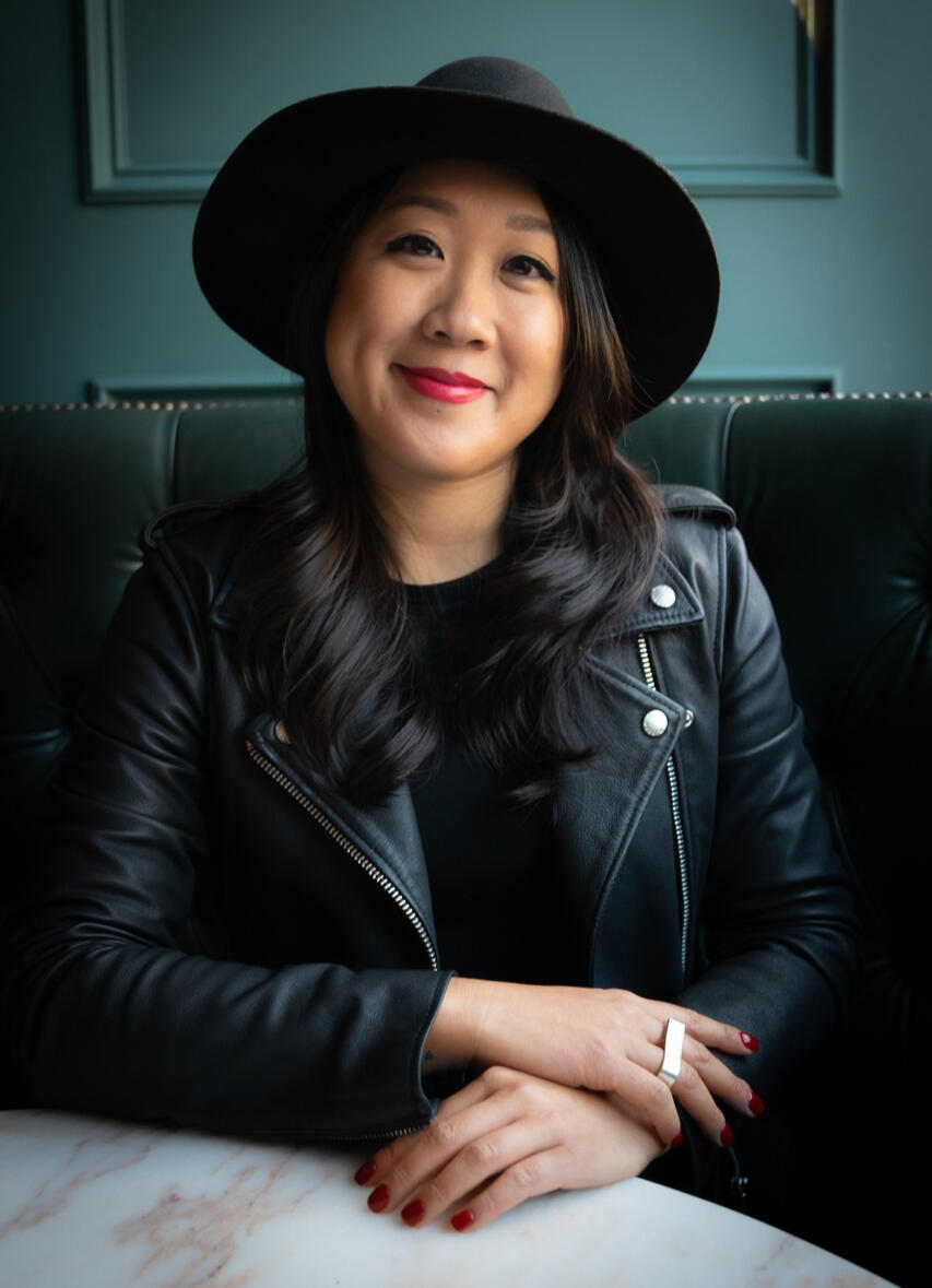 Kris Chau, Small Business Consultant, sits at a table with her hands crossed. She's wearing a black fedora and a black leather jacket. Her nails are painted red, and she has a silver bar ring. She's smiling with red lipstick. She helps service-based entrep